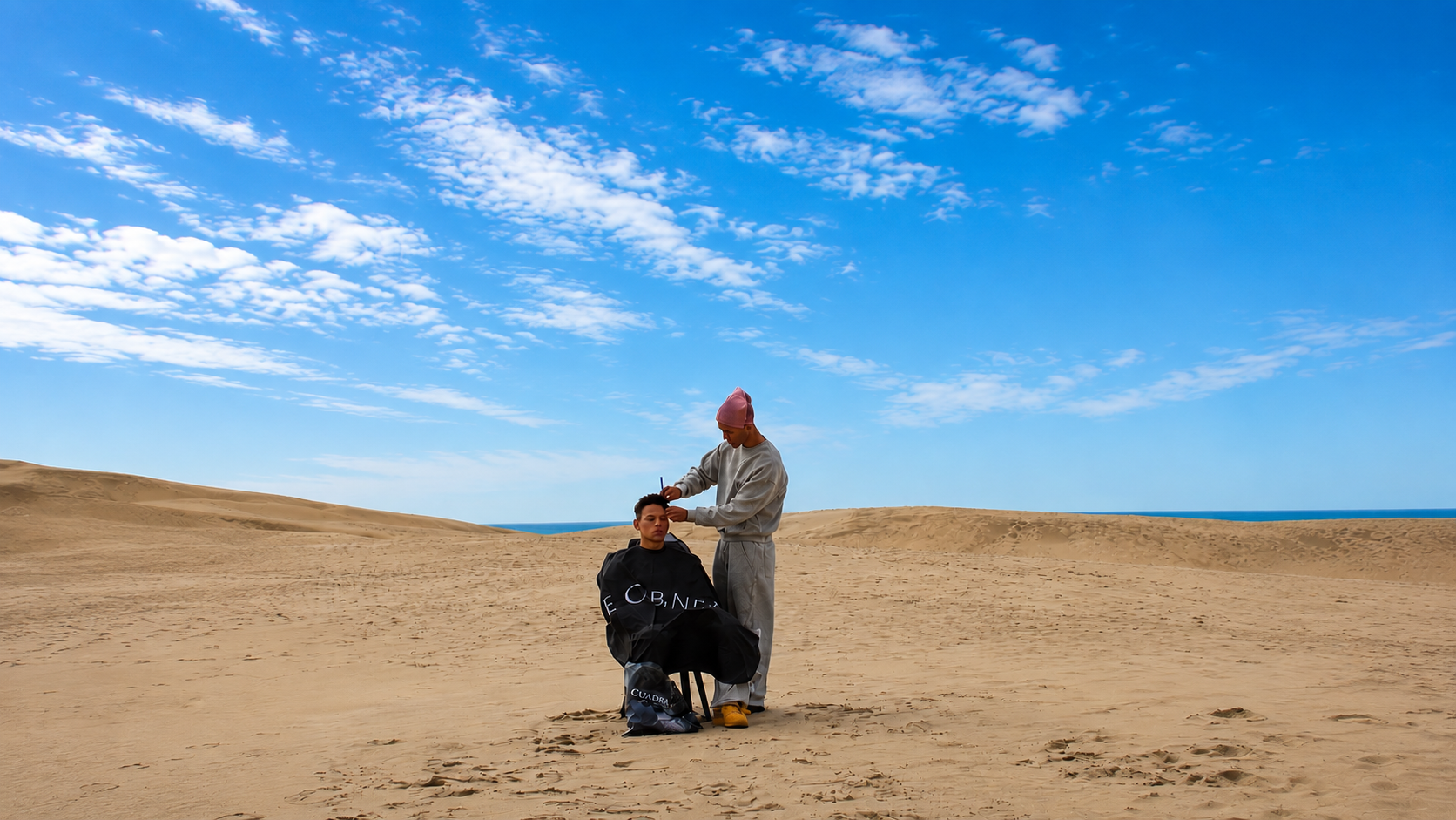 Mehdi coiffe un client dans un paysage de sable sous un ciel bleu