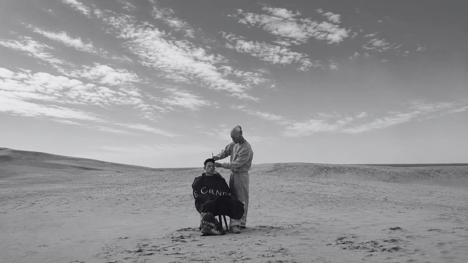 Mehdi coiffant un client dans les dunes, photographie en noir et blanc